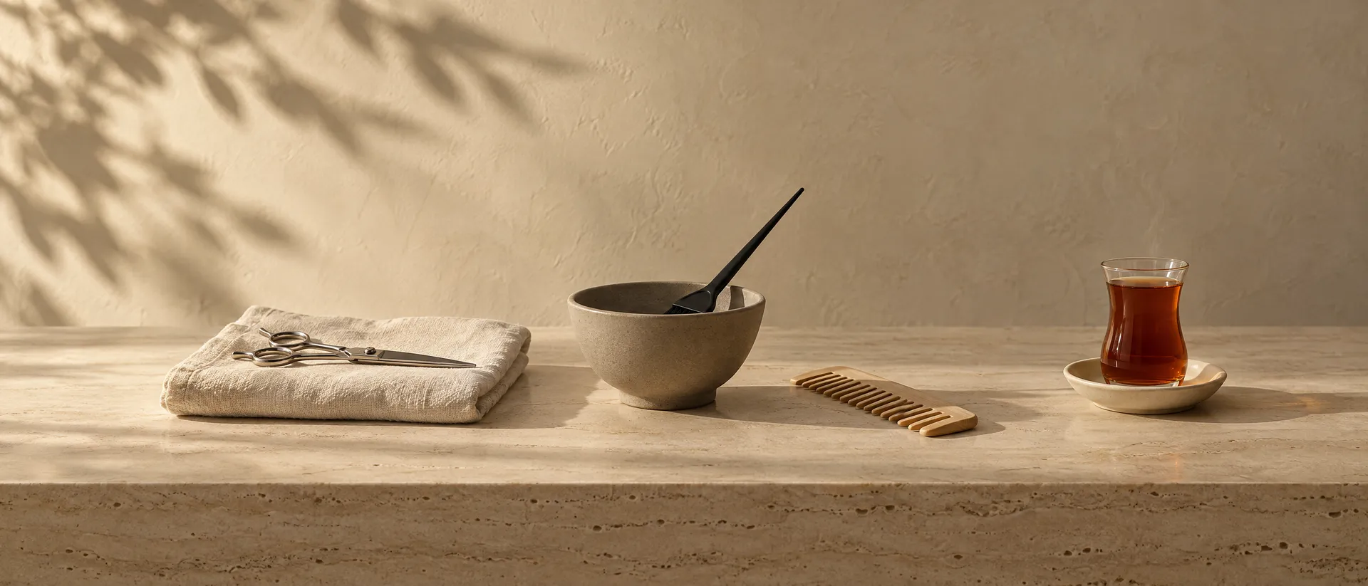 A hairdresser's work surface in morning light: steel shears on a folded linen towel, a stoneware colour bowl and brush, a wooden comb and a glass of Persian tea on travertine.